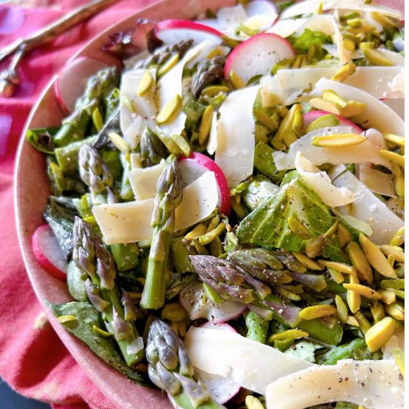 Fresh asparagus salad with radishes, cheese shavings, and pistachios on a pink plate.