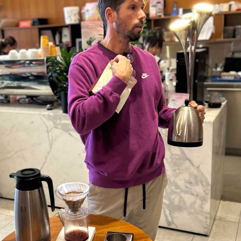 Man in purple sweater holding kettle near coffee setup on small table.