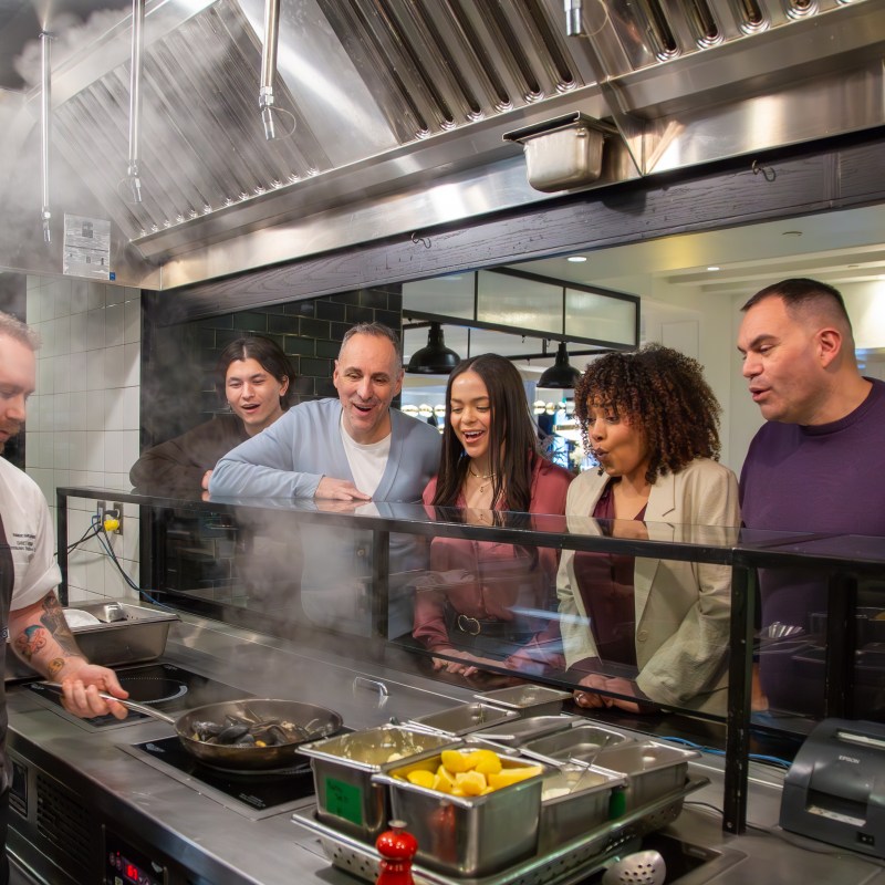 Chef cooking while five people watch through a glass partition in a kitchen.