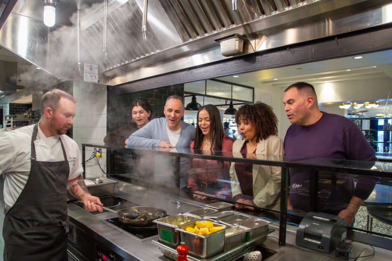 Chef cooking while five people watch through a glass partition in a kitchen.