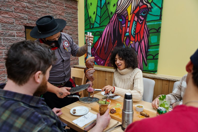 Waiter serves grilled meat on skewer to smiling diners at table with colorful mural background.