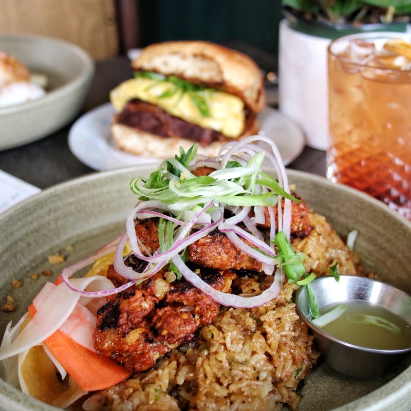 Close-up of rice and grilled meat dish with herbs, pickled vegetables, and a drink in the background.