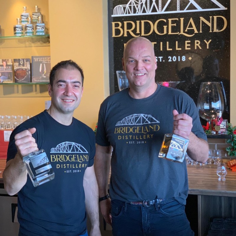 Two men smiling, holding bottles at Bridgeland Distillery.