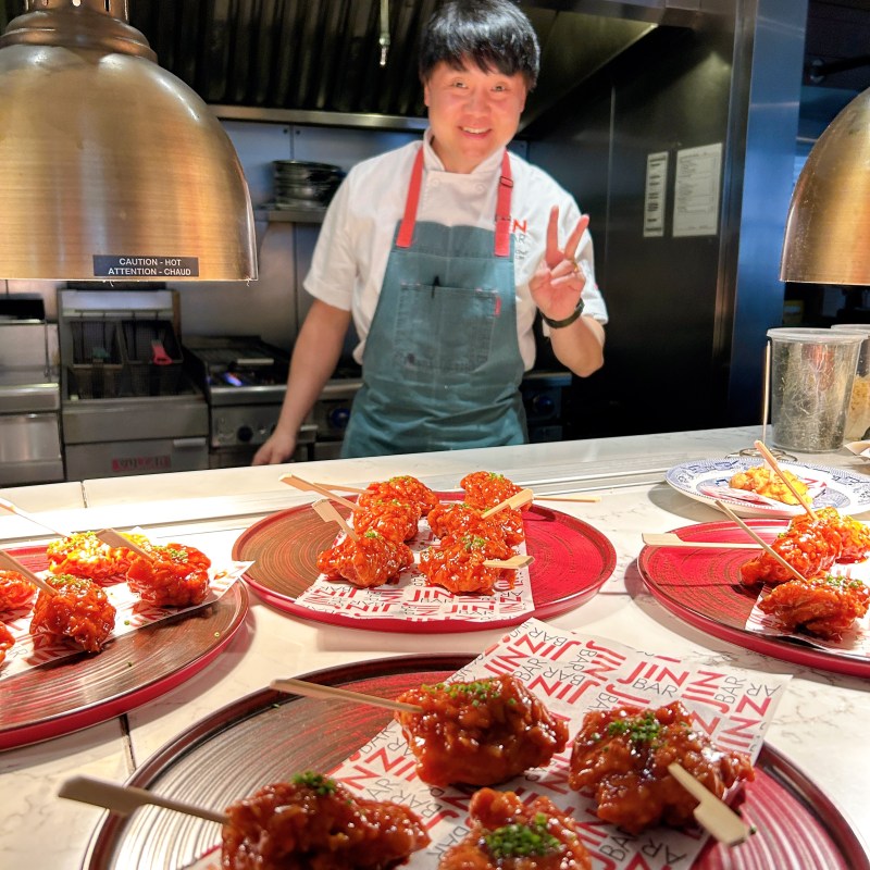 Chef posing with peace sign behind plates of glazed chicken skewers.
